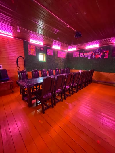 The image shows an interior scene in what appears to be a room with unique decor. Walls are wooden, illuminated with neon lights creating a pinkish-red hue. There is artificial turf on a portion of the wall adorned with framed pictures or posters. A line of stacked, dark-colored wooden chairs is placed near the transition between the wooden wall and the turf. A window is visible on the turf-covered part of the wall. The scene is likely indoors, but there are no people visible in the image. There is no indication of the city of Картмазово, Russia. The focus of the image is on the interior decor and the arrangement of the chairs.