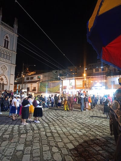 This image depicts a nighttime scene in the city of El Cisne, Ecuador. A crowd of people has gathered in a square paved with cobblestones. To the left, a Gothic-style church with a visible rose window is illuminated. In the background, buildings with warm lighting create a backdrop to the square. Some individuals are dressed in traditional clothing, while others are costumed, possibly for a local festival or celebration. A flag with yellow, blue, and red stripes is partially visible on the right. The atmosphere is active, with people standing, sitting, and interacting. Overhead power lines are visible against the dark sky.