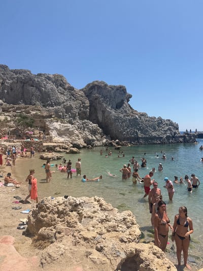 A crowded, rocky beach in Lindos, Greece, under a clear blue sky. Numerous people, predominantly adults, are engaged in various beach activities. Many are wading in the shallow, clear turquoise water, while others are swimming or floating on inflatable devices. Some individuals are sunbathing on the sandy shore, and a few are gathered near the rocky cliffs that dominate the background. The terrain is characterized by large, rough, light-colored rocks and a sandy beach area. The water appears calm and inviting. The overall scene depicts a popular tourist destination on a sunny day.