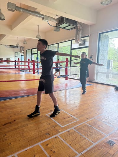 In Cherna Mesta, Bulgaria, three young individuals are engaged in a training session within an indoor gymnasium, possibly a boxing or wrestling facility. The room features a polished wooden floor marked with white tape, forming squares that likely delineate training areas or drills. In the foreground, a boy with dark hair wears a black t-shirt and shorts, both bearing a "Blood Rich" logo, and specialized black and gold boxing shoes. He is actively moving, with his arms extended. Behind him, another boy in a dark t-shirt and shorts stands further back, also appearing to be training. To the right, a third individual, dressed in a green tracksuit with "Los Angeles" visible on the sleeve, stands with arms outstretched, facing the opposite direction from the foreground subject. The background includes a boxing ring with red and yellow ropes and mats, and large windows offering a view of lush green trees. Overhead, industrial ventilation ducts and lighting fixtures are visible on the cream-colored ceiling. The overall scene suggests a focused and active sports training environment.