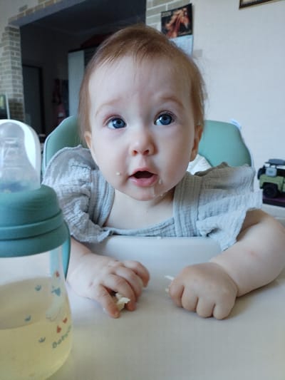 A close-up, eye-level shot shows a baby girl sitting in a high chair with a bottle of milk in the foreground. The baby has blue eyes and fair skin, with a small piece of food on her cheek. She is wearing a grey, ruffled shirt. The background shows a brick wall and a toy car. The scene appears to be in Vinogradny, Russia.