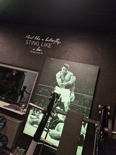 The image displays an indoor fitness area situated in Oberseelbach, Germany. The walls are finished in a dark, textured material, likely black or dark gray. On the upper left wall, a white script reads, "Float like a butterfly, STING LIKE a bee. - Muhammad Ali -". Dominating the right side of the frame is a large, monochrome artwork of boxer Muhammad Ali, depicted shirtless, wearing boxing shorts and gloves, in a fighting stance within a boxing ring. Below this main image, two smaller, partially obscured monochrome images show boxers on the ground. A black weight bench with a padded surface and an integrated bar rack occupies the foreground, partially obstructing the lower portion of the artwork. To the left, a window or glass partition reflects internal elements, including an indistinct figure and additional equipment. A white shelf below this window holds a folded dark blue towel and a clear plastic water bottle with a blue cap. A radiator is visible beneath the shelf. In the lower-left corner, several weight plates with blue accents are present on the floor or a rack.
