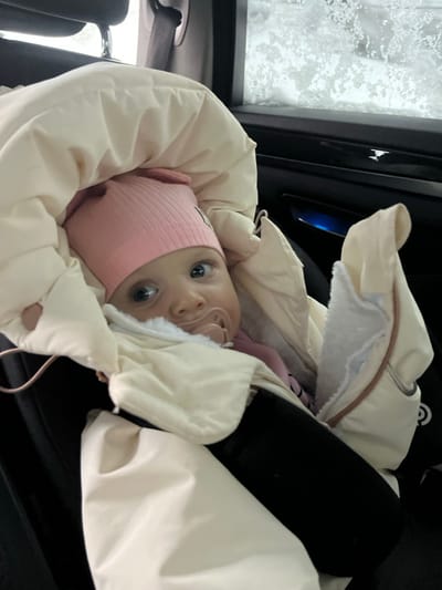 The scene is located in Vysokovo, Russia, inside the rear passenger compartment of a vehicle. A single infant is depicted, secured in a black car seat or pram attachment, mostly enveloped within a cream-colored, padded sleeping bag or winter cover with a fuzzy white interior lining visible around the face. The infant wears a pink ribbed beanie with small ear-like protrusions and a white emblem on the forehead, and has light blue eyes and fair skin. A pink pacifier is situated in the infant's mouth. The infant's legs are covered by black material, possibly leggings or part of an outfit. A brown cord is visible on the left side of the sleeping bag. The infant is looking directly forward, towards the viewer, with an alert expression. The vehicle interior features dark upholstery and door panels; a portion of a headrest is visible in the upper left, and a blue ambient light detail is present on the right-side door panel. Outside the window on the right, a blurred, snowy, or icy landscape is visible, indicating cold weather conditions. The window glass exhibits visible frost or ice accumulation.