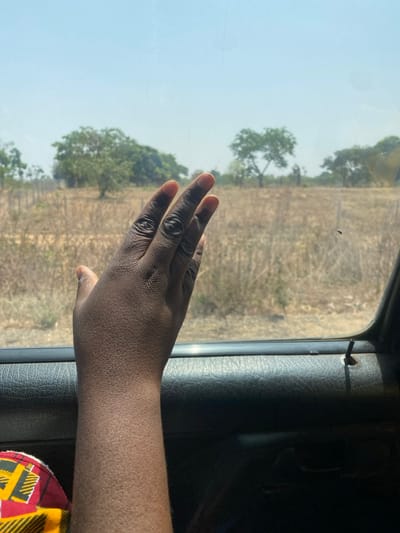 The image depicts a forensic view from inside a vehicle looking out a window onto an arid landscape in Unguwar Gaya, Nigeria, under a clear sky. In the foreground, the dark-skinned forearm and hand of a person are prominently displayed on the left, with fingers slightly splayed and extended towards the window. A fragment of patterned fabric, featuring red, yellow, and black geometric designs, is visible in the bottom left corner. The bottom and right portions of the frame are occupied by a dark, textured vehicle door panel, showing what appears to be a recess or attachment point near the right edge. Outside the window, a dry, brownish field with sparse vegetation and bare ground extends into the midground. A rudimentary fence line, comprising vertical posts and horizontal elements, traverses the field horizontally. Several trees with green foliage are scattered throughout the landscape. A small insect, resembling a fly, is present on the windowpane in the mid-right. The background features a light blue, cloudless sky. The scene is illuminated by bright sunlight.