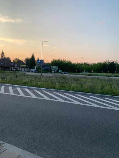 This is a low-angle, outdoor, eye-level, full shot of a road and surrounding environment in Nowodworce, Poland, likely during early morning or late evening due to the soft, warm lighting. In the foreground, a paved road curves into view, marked with white diagonal lines indicating a lane or traffic direction. To the left and stretching into the mid-ground is a grassy area with tall weeds and scattered wildflowers. Beyond the grassy area, a street is visible with several cars, buildings, and signage. On the left, a two-story building with a dark roof and brown siding is present, resembling a residential or commercial structure. Adjacent to it is a smaller building with a yellow facade, likely a shop, featuring display windows. A street lamp stands near the shop. Further back, several trees form a dense line along the horizon, with more trees and foliage interspersed with signs. One sign, clearly legible, reads "DOM" with an arrow, and another smaller sign indicates "Portofino". A few more street lamps are visible in the mid-ground and background. The sky above is a gradient of soft orange and light blue, with subtle cloud formations. The overall scene depicts a suburban or semi-rural intersection with traffic infrastructure and modest buildings.