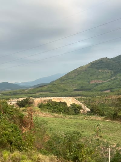 The image presents a view of a landscape in Aê Thi, Vietnam. There is a series of mountains receding into the background under a cloudy sky, with electrical power lines extending across the sky in the upper left. A stone wall runs parallel to the mountains on the right, overgrown with vegetation. There are areas of green grass and dense shrubbery in the foreground. The scene is oriented vertically.