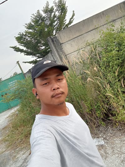 A man wearing a black baseball cap and a grey t-shirt takes a selfie outdoors. He is positioned in the foreground, with a concrete wall and overgrown vegetation behind him. A green metal gate with writing is visible on the left side of the image. The location is specified as Teluk Kenidai, Indonesia. The man has a mustache and is wearing an earpiece in his left ear. The sky is overcast.