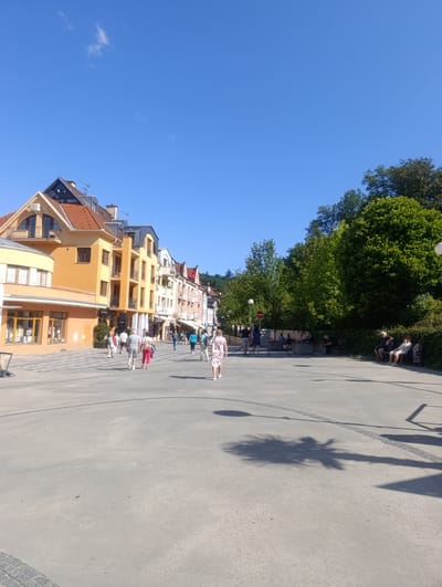 The image depicts a sunny day in Luhačovice, Czechia, likely on a pedestrianized street lined with colorful buildings and lush greenery. Several individuals are seen walking on the paved walkway. Some are seated on benches, enjoying the weather. The architecture suggests a European town. The scene is bright and airy with a clear blue sky overhead.