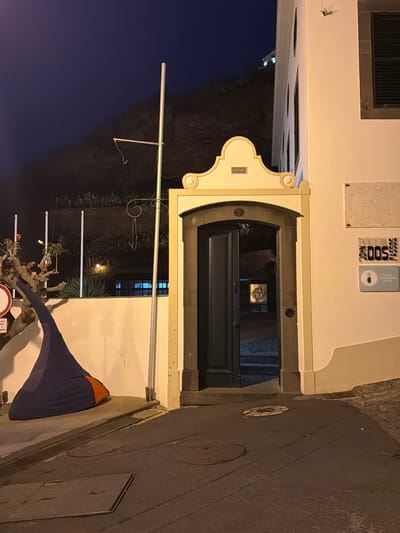 The image captures a night view in Ponta do Sol, Portugal, focused on the entrance to the John Dos Passos Cultural Centre. The building's facade is off-white, with a dark window on the upper left and memorial plaques above the entrance. The entrance is an arched doorway, leading into a dark passage. The street is to the right, showing dark asphalt with manhole covers. A blue and orange object, possibly a sunshade or covering, is positioned in front of the building. A lamppost and some vegetation are visible in the foreground. The sky is dark in the background, indicating the night setting.