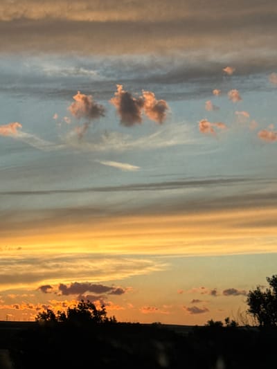 The image depicts a sunset over a rural landscape, likely in or near Struino, Bulgaria. The sky is filled with layers of clouds painted in hues of orange, yellow, and muted blues and grays. Some of the clouds are tinged with pink, suggesting the light of the setting sun. In the foreground, a dark silhouette of trees and vegetation borders the bottom of the frame, with a faint hint of a horizon line visible beneath the sky. No people or specific man-made structures are clearly identifiable in the scene, emphasizing the natural beauty of the twilight sky and the landscape.