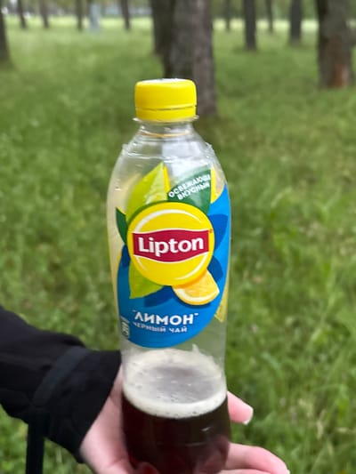 A person's hand holds a bottle of Lipton iced tea with a yellow cap and label featuring a lemon slice and the Lipton logo. The bottle is partially filled with a dark liquid and foam. The background shows a blurry green grassy area with trees, suggesting an outdoor setting. The image is taken in Nevyansk, Russia.