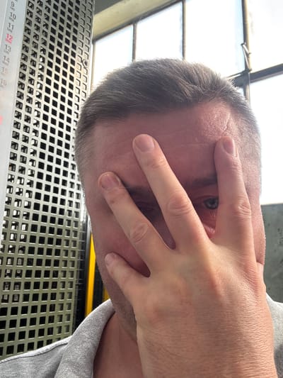 The image shows a close-up of a person's face, partially obscured by their hand. The person has short, dark hair and is wearing a grey shirt. The background includes a metal grate structure with a vertical calendar display showing dates in Italian. The calendar shows the month of May, with days numbered from 9 to 16. The environment appears to be indoors, with large windows visible in the background. No specific location in Mejaniga, Italy, is identifiable from the image.