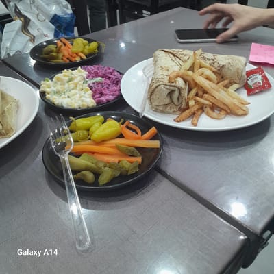 The image shows a meal consisting of shawarma wraps with french fries and a side of pickled vegetables and salad, plated on a metal table. The food is served on white and black plates. A small packet of Heinz ketchup is visible near the shawarma. A person's hand and a phone are near the plate of shawarma. Pink paper with writing is also near the shawarma. The environment appears to be a casual dining setting, likely in a restaurant. The text "Galaxy A14" is watermarked on the image. There are no obvious signs indicating the precise location as Nuseirat.