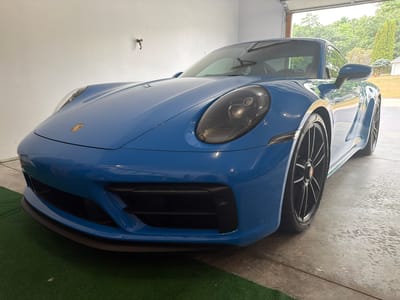 A blue Porsche 911 GT3 is parked in a garage in Wisconsin Dells, Wisconsin, United States of America. The car is facing towards the viewer, with its front end slightly angled to the right. The garage appears to be a residential garage, with a concrete floor and white walls. The garage door is open, revealing a suburban backyard with trees and a house in the background. The car is clean and appears to be in good condition. The lighting in the garage is a mixture of natural light from the open garage door and artificial light from overhead fixtures. The floor of the garage is wet, suggesting it may have recently rained or been washed. The car has black rims and red brake calipers. The car's headlights are on, and the interior of the car is visible through the windshield. A small green mat is positioned in front of the car, on the garage floor.