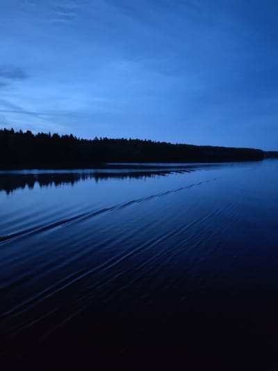 The image shows a dark, tranquil body of water at dusk, with a dense line of trees silhouetted against the deep blue sky along the far bank. The water is smooth, with subtle ripples extending from the foreground towards the tree line, suggesting movement, possibly from a boat or the wind. The reflections of the trees are faintly visible on the water's surface. The scene is set in Yakunina Gora, Russia, a location known for its natural beauty.