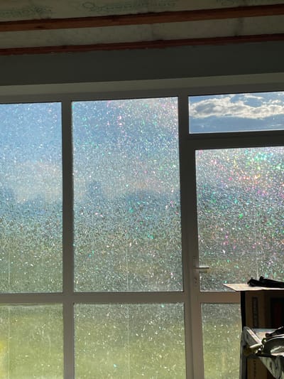 The image shows a window with patterned privacy film, which refracts light into a rainbow of colors. Through the top left pane, a bright blue sky with fluffy white clouds is visible, suggesting daytime and clear weather. The rest of the window panes are covered with the shimmering film, obscuring the view outside but hinting at a green, possibly natural, landscape beyond the right-hand panes.
To the left of the window, construction materials are visible, including wooden beams and insulation that appears to be covered with a patterned material. This suggests the window is part of a building undergoing renovation or construction.
At the top right of the image, above the window, a partial view of some boxes and equipment is visible. One box has text printed on it: "80 V (PRO)". The overall impression is of an indoor space looking out through a specially treated window, with evidence of ongoing work. The light filtering through the patterned film creates a visually dynamic and somewhat abstract effect. No people are visible in the image. The context of Izborsk, Russia, is provided but not directly evident from the visual content of the image itself, other than perhaps implying a Northern latitude for the sky.