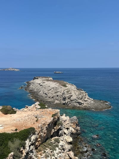 The image shows a scenic coastal landscape in Rizokarpaso, Northern Cyprus. The scene is dominated by a clear, bright blue sky and a vast expanse of deep blue ocean. In the foreground, a rocky coastline with rugged cliffs juts out into the sea. The rocks are varied in color, ranging from light gray to brown and are interspersed with patches of green vegetation, including bushes and shrubs. The water near the shore is a striking turquoise, revealing the rocky seabed beneath its surface.
Further out in the sea, several small, rocky islands are visible. The largest of these islands, situated in the mid-ground, is a prominent feature with a flat, rocky surface and sparse vegetation. In the distance, on the horizon, more islands can be seen, appearing as small, rugged shapes against the blue of the sea. A small, dark object, possibly a boat or a structure, is visible on the water in the distance. The overall impression is one of a remote and untouched natural environment. There are no people or animals visible in the image.