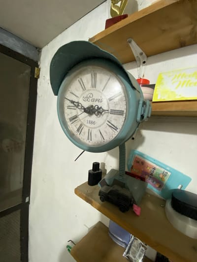 The image shows a vintage-style teal clock on a desk with several items. The clock has Roman numerals and the word "Paris" on its face, with the year "1886" at the bottom. The clock is mounted on a flexible metal stand and has a protective visor. Next to the clock, a small black toy car is visible. A light blue plastic holder with some printed items is also on the desk. The background includes a white wall, a wooden shelf above, and a doorframe to the left. The setting appears to be an indoor storage area or a room with shelves, possibly in San Jose del Monte, Philippines.
