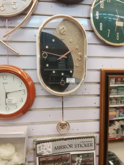 The image depicts a retail display of clocks and decorative items on a white slatwall in a store located in Aketu-Oja, Nigeria. In the foreground, a round, wooden-framed clock with a white face and black hands is visible on the left. To its right, an oval-shaped clock with a gold and black glitter face and a gold frame hangs from the wall. This clock has a pendulum with a gold, circular ornament. Further to the right, partially visible, is a portion of a framed mirror reflecting shelves stocked with various items, likely in the background of the store. Above the oval clock, a dark green, round clock with gold numbers and hands is mounted on the slatwall. Below the clocks, there is a display of what appears to be a "Mirror Sticker" product with text indicating its size as "LX Size: 37x31cm". To the far left, a white box with white flowers is also part of the display. The overall environment suggests a shop selling home decor or accessories.