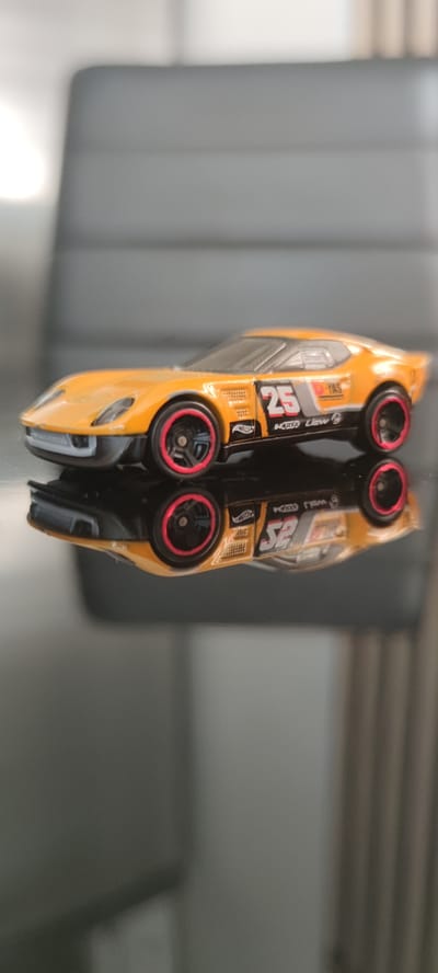The media file shows a close-up, side profile of a miniature orange toy sports car, likely a Hot Wheels model. The car is positioned on a reflective, dark surface, creating a mirror image below it. The car itself features racing decals, including the number "25" prominently displayed on the side, along with other sponsor logos and text, some of which are too blurry to read clearly. The wheels have black rims with a red outer ring. The background is out of focus, appearing as soft grey and white tones, suggesting an indoor setting with diffuse lighting. The overall impression is that of a still-life photograph focusing on the detail and design of the toy car. There are no people visible in the frame. The time of day and weather are not discernible from the image. The location context of Liebenburg, Germany, is not visually apparent in the media file itself.