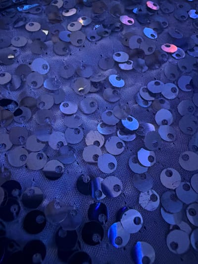 This image depicts a close-up view of a surface in Long Beach, United States of America, densely covered with numerous small, circular, metallic-sheen discs, consistent with sequins. Each disc measures approximately 0.5 to 1 centimeter in diameter and features a central perforation. The discs are affixed to a blue textile fabric, which presents a subtle woven texture. Illumination is primarily blue, causing the discs to reflect various shades of blue light, with occasional iridescent reflections of pink and purple. No individuals, animals, actions, or interactions are visible within the frame. The scene is static.