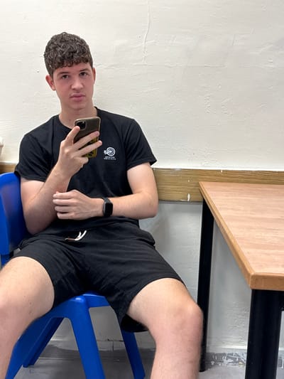 A young male with curly brown hair is depicted seated in an indoor setting in Ness Ziona, Israel. He wears a black short-sleeved t-shirt featuring a white circular logo with Hebrew text on the left chest, black shorts with visible white drawstrings, and a dark smartwatch on his left wrist. He holds a dark-colored smartphone, equipped with a yellow case, in his right hand, with his thumb positioned over the screen. His left hand rests on his right forearm. He is seated on a blue plastic chair. To his right, a table with a light brown top and dark legs is visible. The background consists of a textured, light-colored wall, a wooden trim along the lower section showing white paint or wear, and a light gray floor.