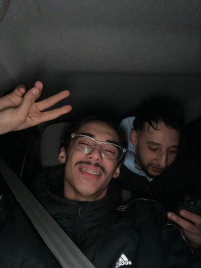 Two young men are in a car. One is asleep in the top right corner, with his eyes closed and head resting on a light blue fabric. The other man is in the foreground, awake and giving the peace sign. He has glasses, braces, and a mustache. He is wearing a black jacket with the Adidas logo on the sleeve. A seatbelt crosses diagonally in front of him. The background appears to be a car interior.