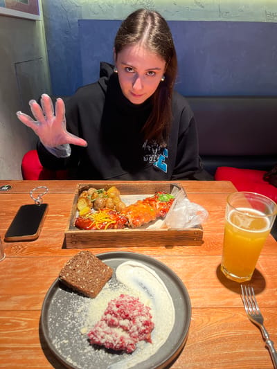 The image depicts a female individual seated at a wooden table in an indoor restaurant setting in Родниковское сельское поселение, Ukraine. The subject, a young woman with long dark hair, is positioned centrally and looks directly at the camera. She wears a black hoodie with a light blue and white graphic. Her right hand, wearing a clear disposable plastic glove, is raised and open, facing the camera.
On the wooden table in front of her, various food items and personal belongings are arranged. A rectangular wooden tray holds roasted baby potatoes and two pieces of meat, possibly ribs, glazed with different sauces and garnished. One piece has yellow string-like garnish, and the other has green chopped garnish. Adjacent to the tray, a dark gray plate contains a portion of minced red meat, likely steak tartare, topped with grated white cheese, a dollop of white cream or sauce, and a slice of dark seeded bread. A tall glass filled with an amber beverage, consistent with beer, sits to the right of the food. A silver fork is placed nearby. A dark smartphone with a gold-colored frame and a sparkling ring or bracelet on top lies horizontally on the table to the left.
The background features a textured wall, split horizontally into an upper section of muted blue-green and a lower section of darker blue-gray. A red cushioned booth seat is visible behind the table. The lighting is dim, with prominent illumination on the table and the subject.