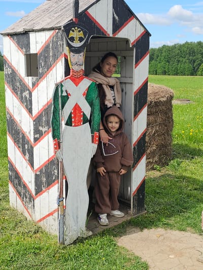 Two individuals, an adult female and a young child, are positioned within a wooden structure resembling a guardhouse. The structure is painted with black and white chevrons interspersed with red stripes. On the side of the structure, a painted depiction of a historical soldier is visible. The adult is lying behind the child who is in the foreground, each looking toward the camera. The environment includes a field with grass and a haystack visible in the background. The photograph was taken in the Borodinskogo Muzeya, Russia.
