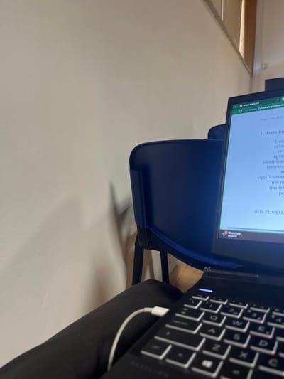 An individual is situated indoors in Gandra, Portugal, with a black laptop resting on their lap, which is clad in dark pants. A white cable is connected to the left side of the laptop. The laptop screen is illuminated, displaying a document titled "Artigo 1 intro.pdf" with Portuguese text, including "Palavras-chave" and "1. Introdução." In the immediate background, a dark blue plastic chair with a black metal frame is partially visible. Further back, a light-colored wall occupies the left side of the frame, exhibiting shadows from an unseen object. In the upper right background, a window with horizontal blinds is partially visible.