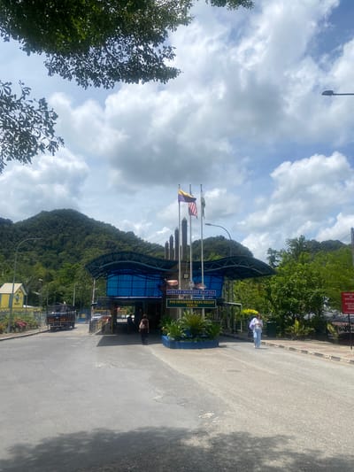 The image shows the Jabatan Imigresen Malaysia immigration post in Wang Kelian, Perlis, Malaysia. The entrance to the post is marked by a sign displaying the name. Flags fly above the covered entrance area. A person is walking on the road near the entrance. Mountains covered in lush greenery rise in the background. The sky is partly cloudy. A section of a road is visible in the foreground. There is a lamppost in the background near the mountain.
