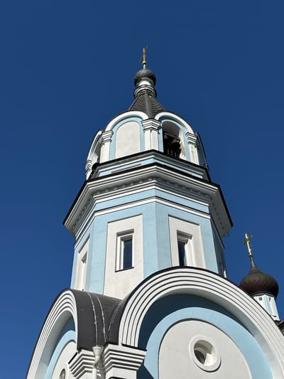 This is a low-angle, close-up, exterior photograph of a light blue and white domed building against a clear, deep blue sky. The building appears to be an Orthodox church, evidenced by the onion domes and golden crosses topping them. The architecture features multiple layers of decorative cornices and arched elements. In the foreground, a section of a dark gray, curved roof with white decorative trim is visible. Above this, a taller section of the building painted light blue and white has rectangular windows and decorative moldings. Further up, a bell tower section is visible with a dark gray, domed roof topped with a golden cross. The building is illuminated by direct sunlight, creating distinct shadows and highlights. The scene is devoid of people or any other visible objects or activities. The location is identified as St. Ozerki, Russia.