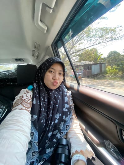 A woman in a hijab and patterned blouse is taking a selfie from the passenger seat of a vehicle. She is making a kissing face and her left arm is visible, adorned with a white watch. The vehicle's interior is dimly lit, with a window on the right showing a sunlit outdoor scene of trees and a small structure resembling a roadside stall or shop in Mandar Sari, Indonesia. The vehicle's ceiling features a handle and an air vent. The woman's right side is obscured by the seat and other objects.