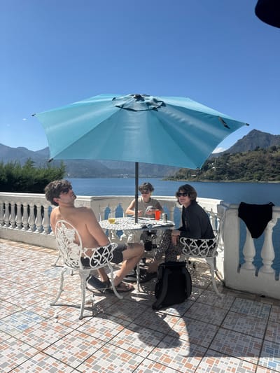 Three individuals are seated at a white metal table on a tiled patio overlooking a body of water and distant mountains. A large blue umbrella provides shade. A person without a shirt sits facing the viewer, while the others are seated at the table facing away. A black garment hangs on the white concrete railing. A black bag sits on the tiled ground. The sky is clear blue.