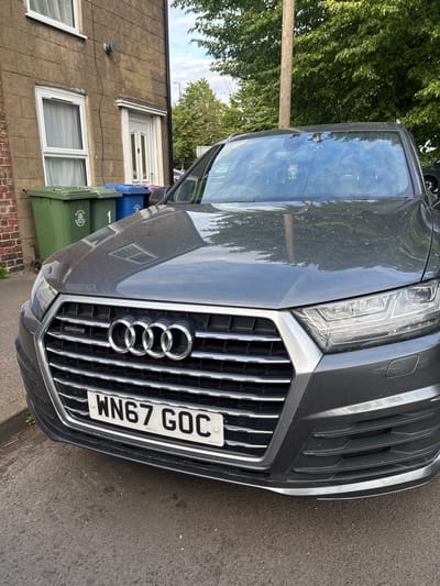 A dark grey Audi Q7, registered with UK license plate WN67 GOC, is parked on an asphalt road in front of a residential building. The vehicle exhibits minor dust accumulation on its hood and windshield, and a slight scuff is present on the lower passenger side of the front bumper. The "quattro" badge is visible on the vehicle's grille.

To the left of the vehicle, a two-story building constructed of light brown, horizontally coursed brick or rendered material stands. It features two white-framed windows, one on the upper level and one on the lower level, both fitted with internal curtains. A white-framed front door with multiple glass panels is located to the right of the lower window. Above the front door, a circular external vent is affixed to the wall, with a black cable running vertically downwards.

Three refuse bins are positioned in front of the building: two green and one blue. The green bins bear the visible inscription "BOSTON BOROUGH COUNCIL" and a civic crest; one green bin is marked with the number "1". The blue bin also appears to have a similar municipal logo. The presence of the "BOSTON BOROUGH COUNCIL" branding identifies the location as Boston, United Kingdom.

The background features lush green deciduous trees under a partially cloudy sky, with a wooden utility pole visible behind the vehicle's windshield. The scene is illuminated by natural daylight.
