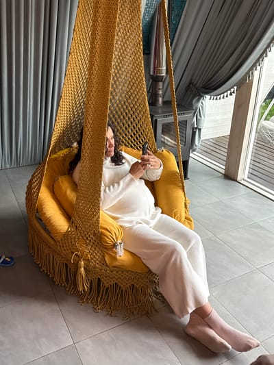 A woman lies in a hanging chair indoors. She is wearing a white sweater and pants, and pink socks. She is holding a black smartphone in her hands. The chair has yellow cushions and a macrame-style weave with fringe. To the left of the chair are gray curtains, a chrome object, and a gray cabinet. A window is in the background. The floor is gray tile. A child's blue flip-flop is visible in the lower right corner. The location is indoors and not identifiable as Чёрное, Russia.