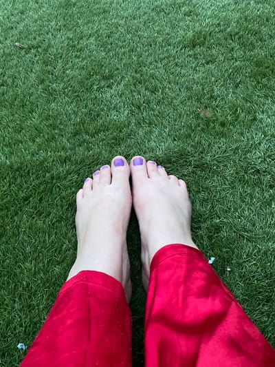 Bare feet with purple toenails are on artificial green grass next to the bottom portion of red pants. The scene appears to be outdoors, but a specific location in Sint-Baafs-Vijve, Belgium, cannot be determined from the image. No actions or interactions are visible.