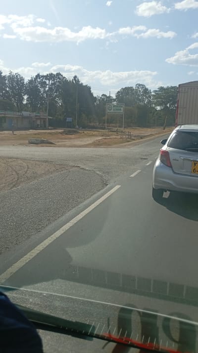 A view from a vehicle, showing a gray car on the right side of the road with license plate KC 64. The road has a marked white line, indicating lanes. A truck is partly visible next to the car. In the background, there is an intersection with a sign that says "WOBURN SCHOOL." The sky is partly cloudy, and trees line the distant horizon. The road surface appears worn and uneven.
