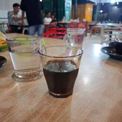 The image depicts a close-up view of two glasses on a wooden table, with a partially visible person in the background. The glass in the foreground contains a dark liquid, likely coffee, and the glass to its left contains a clear liquid, likely water. The table has a light brown, marbled surface. In the background, a person wearing a dark shirt and blue jeans is visible, appearing to be sitting. Further in the background, other blurred figures and red chairs are discernible, suggesting a public setting such as a cafe or restaurant. The lighting is dim, with some bright spots illuminating the table surface. The overall impression is of a casual setting, possibly during an evening or in an indoor environment with artificial lighting. The location is identified as Mesjid Tuha, Indonesia, though this information is external to the visual content of the image itself.