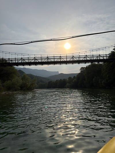 This media file captures a scenic view of a suspension bridge over a body of water, likely a river, near Žunovnica, Bosnia and Herzegovina. The image is oriented vertically.

The primary subjects are the bridge, the water, and the natural landscape. The bridge, made of metal with visible cables and supports, stretches across the frame. Its dark silhouette contrasts with the bright sky and the shimmering water. The water surface is rippled, reflecting the light and the surrounding greenery and sky. Lush green trees line the banks of the water and extend up the slopes of distant, hazy mountains.

The sun is visible in the sky, low on the horizon, suggesting it is either early morning or late afternoon. The sky is partly cloudy, with the sun creating bright flares on the water's surface. The overall atmosphere is serene and peaceful, with a touch of natural grandeur. There are no people visible in the frame, nor any identifiable text. The scene appears to be a calm moment in nature, with the man-made structure of the bridge integrated into the landscape.