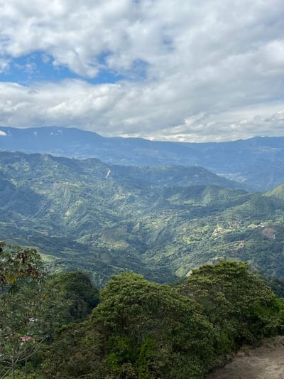 This is an aerial landscape photograph taken in the municipality of Nocaima, Colombia. The image depicts rolling green hills covered in dense vegetation, with layers of mountains stretching into the distance. The sky is partly cloudy, with patches of blue sky visible between white clouds. In the foreground, lush green trees frame the view of the expansive mountain range. Small houses and agricultural plots are visible scattered throughout the valleys and on the hillsides, indicating human habitation and cultivation in this rural setting. The overall impression is one of natural beauty and tranquility.