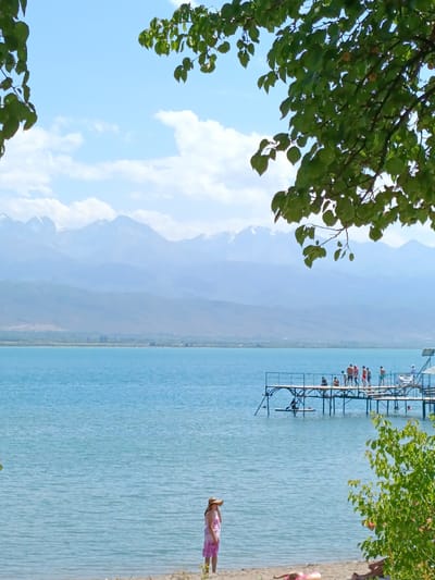 A wide shot captures a serene lake scene with distant, snow-capped mountains under a clear blue sky with scattered white clouds. In the foreground, lush green leaves frame the top of the image, suggesting a tree overhead. A sandy beach runs along the bottom, with a solitary woman wearing a straw hat and a pink dress standing near the water's edge. Further out on the lake, a wooden pier extends from the right side, populated by a group of people, some of whom are standing and others appear to be sitting or interacting. A person is visible on a paddleboard in the water near the pier, and another person is seen swimming. The water itself is a calm, bright blue. The overall impression is a tranquil summer day at a lakeside resort, likely in the vicinity of Kirpichny, Kyrgyzstan, given the mountainous backdrop.
