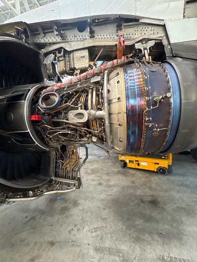 The image shows a disassembled aircraft engine, possibly undergoing maintenance or repair, inside a hangar or workshop. The engine is intricate with visible turbines, piping, and wiring. A yellow aircraft tow tractor is positioned adjacent to the engine. The location in the city of Lautzenhausen, Germany, suggests this is likely at or near the Frankfurt-Hahn Airport (HHN), which is nearby.