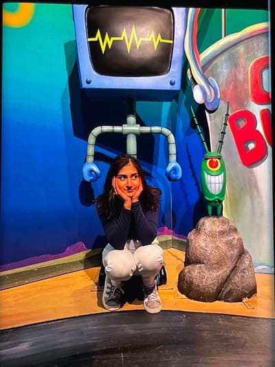 A young woman is seated on a flat, wooden surface in front of a colorful, cartoonish backdrop. The backdrop appears to be inspired by the animated show SpongeBob SquarePants, featuring elements like a large, red, bubbly "B" character and a small, green, one-eyed character with antennae. There's also a large rock-like object and a blue computer monitor displaying a yellow zigzag line. The woman is wearing a dark long-sleeved shirt, light-colored pants, and sneakers. She has her hands on her cheeks, and her expression suggests she is posing for the camera. The lighting is bright, and the overall scene is vibrant and playful. The provided location context of Nijmegen, Netherlands, does not offer specific visual cues within the image itself, but suggests this might be an attraction or event in that area. No visible text is present in the image.