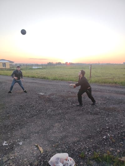 This media file depicts two individuals engaged in a game of catch with a dark-colored ball, which is captured mid-air against a bright, hazy sky. The scene is set outdoors during what appears to be either sunrise or sunset, given the warm, soft light illuminating the horizon with hues of orange and yellow.
One person, wearing a dark jacket and jeans, is positioned on the left side of the frame, in a defensive stance with legs spread apart, seemingly ready to catch or throw the ball. The other individual, dressed in a dark shirt and pants, is on the right, in the process of throwing or catching the ball, with their body turned and arm extended. Both appear to be young men, and their posture suggests an active and playful moment.
The ground is covered in gravel and dirt, with some patches of grass and scattered debris, including a plastic bag and some dry leaves in the foreground. In the background, a rural landscape unfolds. There are grassy fields, some fencing, and a few structures, including a house with a light-colored wall and a dark roof on the far left. Power lines and trees are visible in the distance, hinting at a semi-rural or suburban setting.
The overall atmosphere is relaxed and casual. The bright sky suggests clear or partially cloudy weather. The location context is identified as Rastunovo, Russia. There is no visible text in the image.
