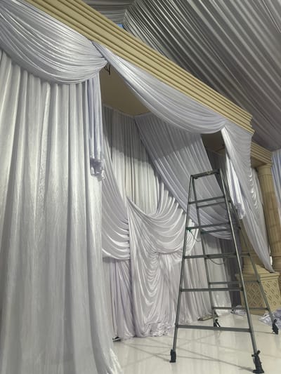 In Langa, Indonesia, a metallic ladder stands on a polished white floor in front of a stage decorated with flowing white drapery. The fabric is artfully draped and bunched, creating a textured and voluminous backdrop. Above the white fabric, a tan-colored decorative structure with horizontal ridges is visible, and a ceiling adorned with gray fabric panels extends upwards. To the right of the ladder, a large, fluted pillar, also in a pale, textured material, supports the overhead structure, and a decorative lattice-like element is visible at its base. The overall scene suggests a venue being prepared or decorated for an event.
