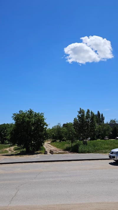 The image depicts a clear blue sky with a cumulus cloud in the upper left quadrant. The right side of the image shows a paved road, with trees and green foliage bordering the road. A portion of a car is visible at the top right corner. The location is consistent with a suburban area or a road on the outskirts of a town. There is nothing present to indicate the specific location of Yerzovka, Russia.