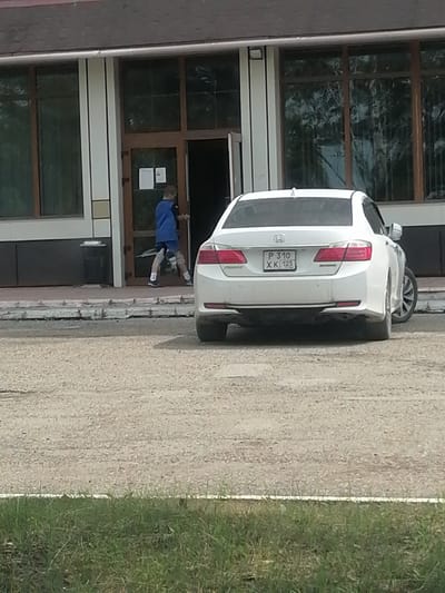 A young person wearing a blue jacket and dark shorts is exiting a building through a double glass door. The person is captured mid-stride, moving from left to right. In front of the building, a white sedan is parked, facing away from the viewer. The license plate of the car is visible and reads "P 310 XK 125". The building appears to be a commercial or public establishment with large windows and a dark roof. The ground in front of the building is a gravel surface, and there is a strip of grass at the bottom of the frame. The scene is set on a cloudy day.