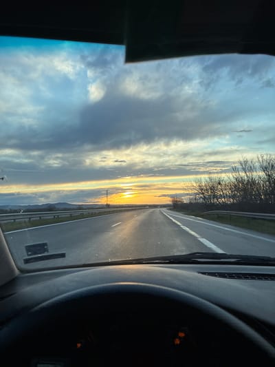 The image depicts a sunset view from inside a vehicle driving on a road in Georgi-Dobrevo, Bulgaria. The sun is setting on the horizon, casting a golden glow on the clouds and the road ahead. The road is a two-lane highway with a white line dividing the lanes. Trees line the side of the road. Part of the dashboard and steering wheel are visible in the foreground. There are no people visible in the frame.