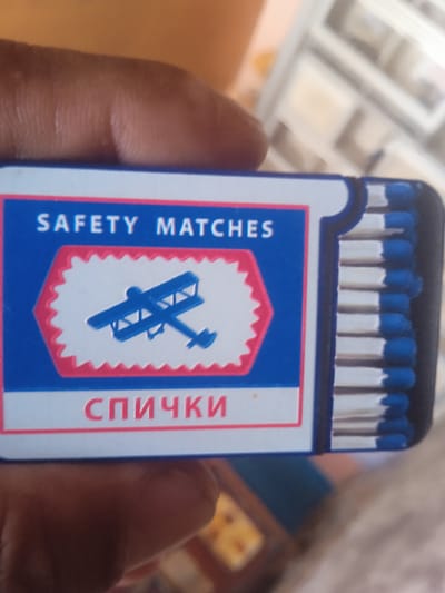 A close-up, angled shot shows a hand holding a box of matches. The box is blue and white with red trim. It has the words "SAFETY MATCHES" printed in white on the blue surface. Below this, there is a white hexagonal shape with a red serrated border, containing a silhouette of a biplane. At the bottom of the box, the word "СПИЧКИ" is printed in red. The matches themselves are visible on the right side of the box, with white shafts and blue heads. The background is blurred, with hints of furniture and possibly other objects. The image appears to have been taken in Kyzyl-Sengir, Kyrgyzstan, though there is no specific visual evidence within the frame to confirm this location.